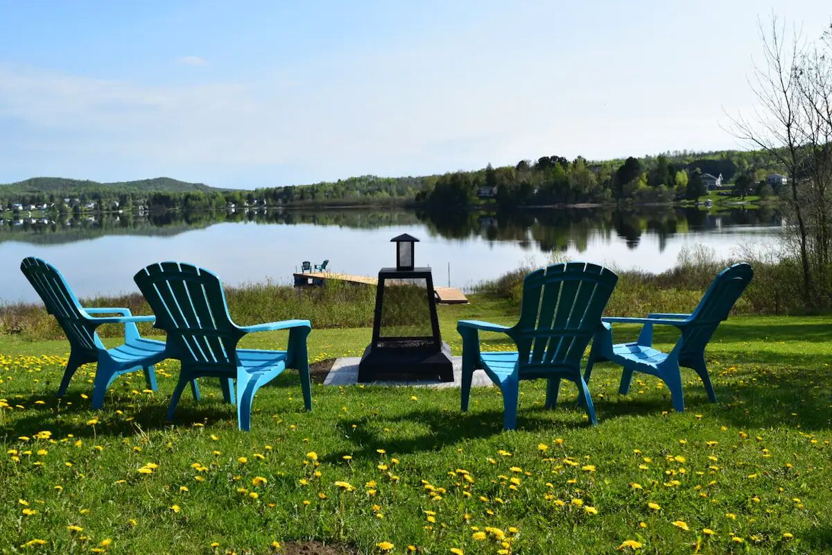 Un cadre idyllique au bord d’un lac, avec trois chaises Adirondack bleues disposées face à un lac paisible, le tout entouré d’herbe verte et de fleurs jaunes.