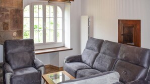 Une pièce avec un mur en pierre, une fenêtre aux cadres blancs, un canapé gris, une table en bois et une porte en bois.