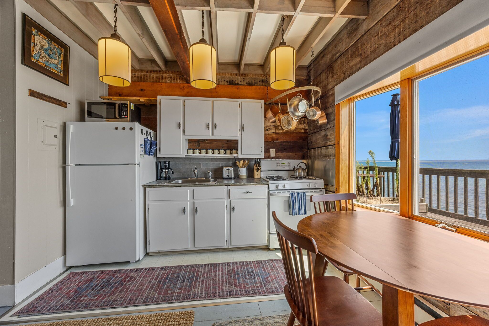 A kitchen with white cabinets, a refrigerator, a stove, and a dining table with chairs. There is a view of the ocean through large windows.
