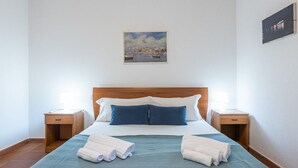 Cama bem arrumada com roupa de cama azul e branca, duas mesas de cabeceira com luminárias, um quadro com cena costeira e piso de azulejos.