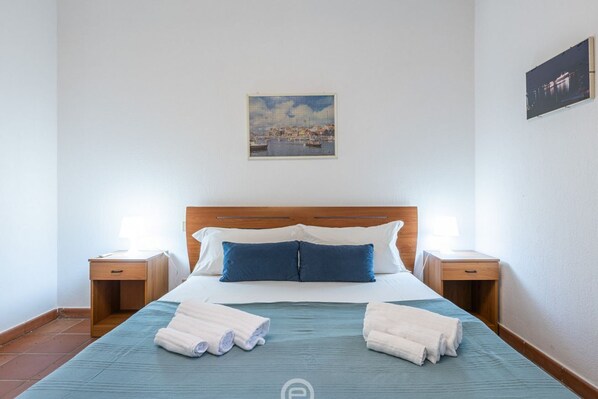 A neatly made bed with blue and white bedding, two bedside tables with lamps, a painting of a coastal scene, and a tiled floor.