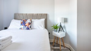 A neatly made bed with white linens and a floral pillow, a bedside table with a lamp and a potted plant, and a white nightstand. - 5* Ultra Luxury, Great Views, Underground Parking (Belfast)