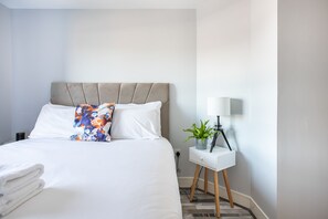 A neatly made bed with white linens and a floral pillow, a bedside table with a lamp and a potted plant, and a white nightstand.
