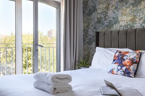 A hotel room with a large window, a neatly made bed with white linens, a colorful pillow, and a plant on the nightstand.