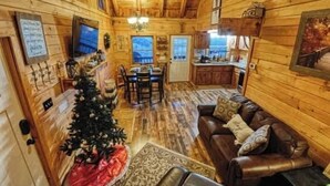 A cozy log cabin interior with a dining area, a living room with a sofa, and a kitchen visible through an open door. - The Nest - Pickle Ball, Firepit, Hot Tub, Mt Views (Sevierville)