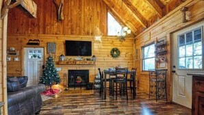 A cozy log cabin interior with a fireplace, wooden walls, a dining area, and a Christmas tree. - The Nest - Pickle Ball, Firepit, Hot Tub, Mt Views (Sevierville)