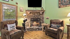 Een gezellige woonkamer met een stenen openhaard, twee leren fauteuils, een houten salontafel en een televisie die boven de openhaard is bevestigd.