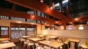 Een restaurant met houten tafels en stoelen, grote ramen en een hoge plafond met zichtbare balken.