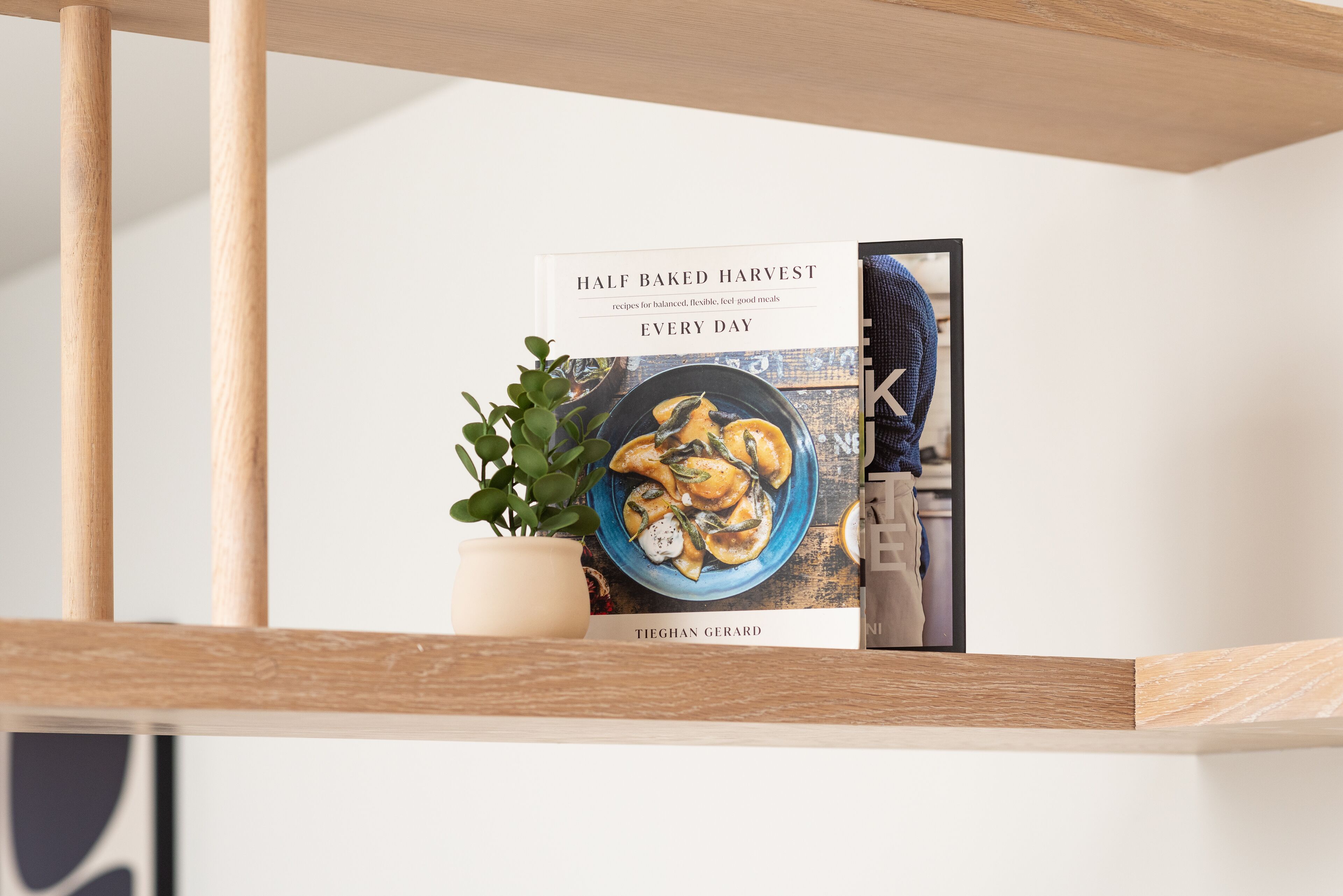 Een boek met de titel 'Half Baked Harvest' van Tieghan Gerard op een plank, met een potplant en een ingelijste foto.