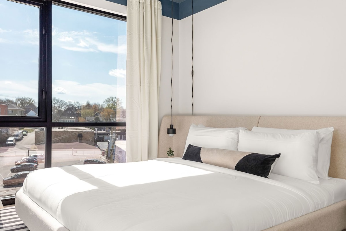 A modern hotel room with a large bed, a view of the street, and a hanging lamp.