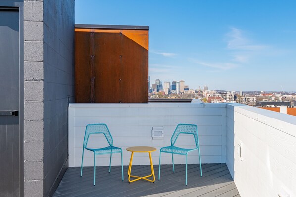 Un balcón con dos sillas turquesas y una mesita redonda, con vistas a un paisaje urbano con edificios modernos.