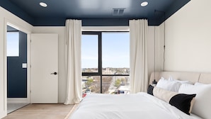 Ein Schlafzimmer mit einem großen Bett, weißen Vorhängen, Blick auf die Stadt und einer dunkelblauen Decke.