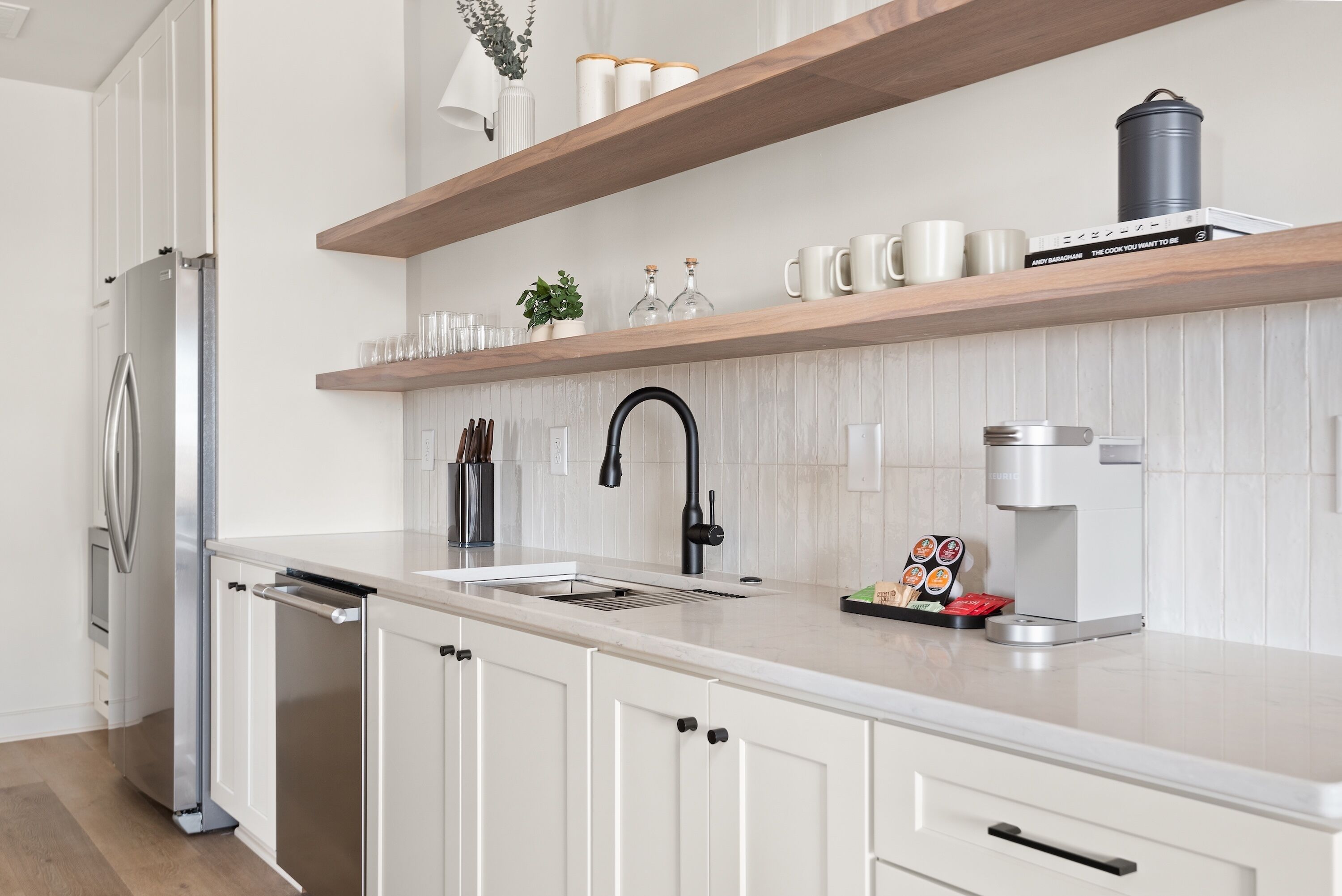A modern kitchen with white cabinetry, a stainless steel refrigerator, a coffee maker, and a shelf with mugs and a book.