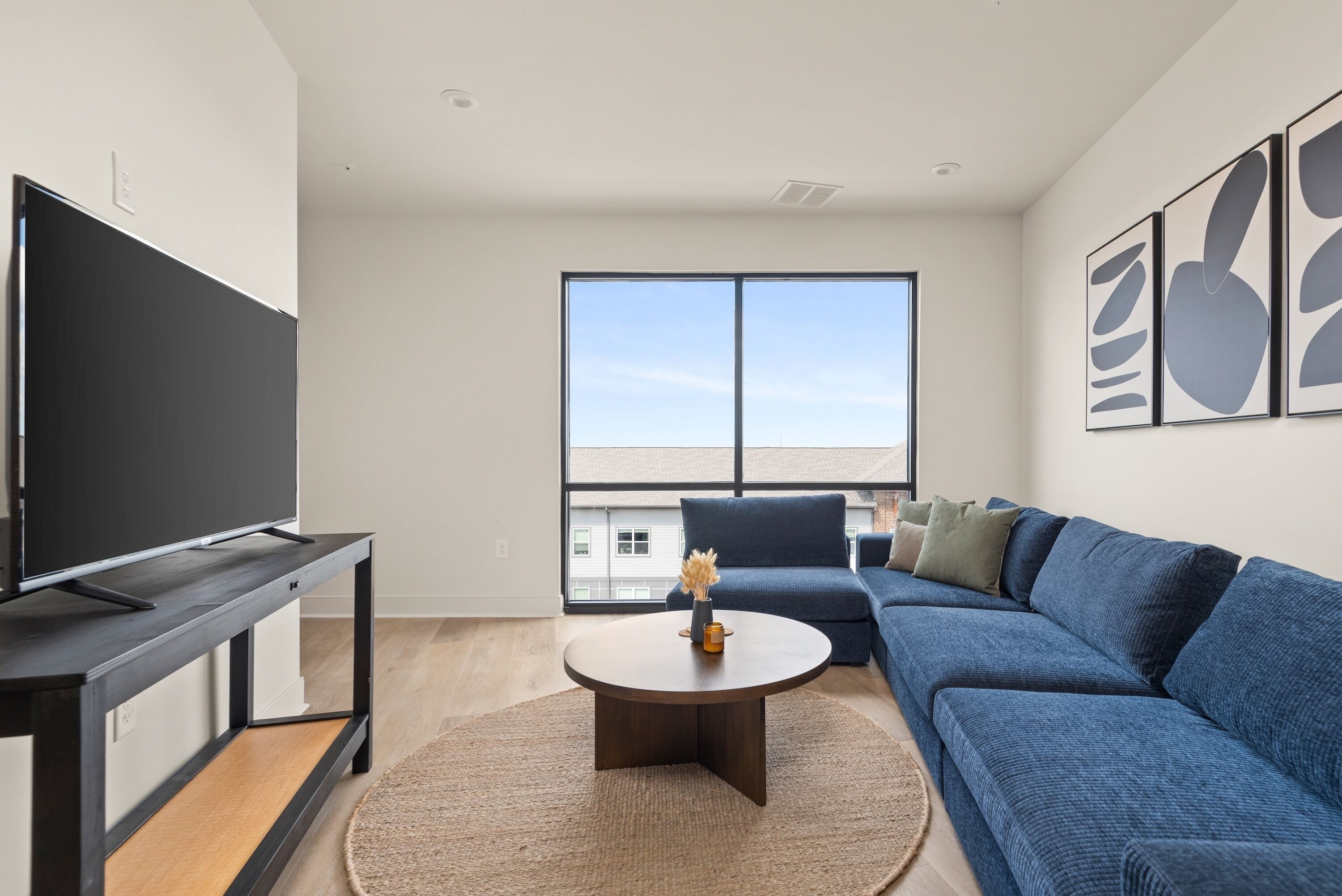 A modern living room with a large flat-screen TV on a black stand, a round coffee table with a vase, a blue sofa, and a large window offering a view of buildings.