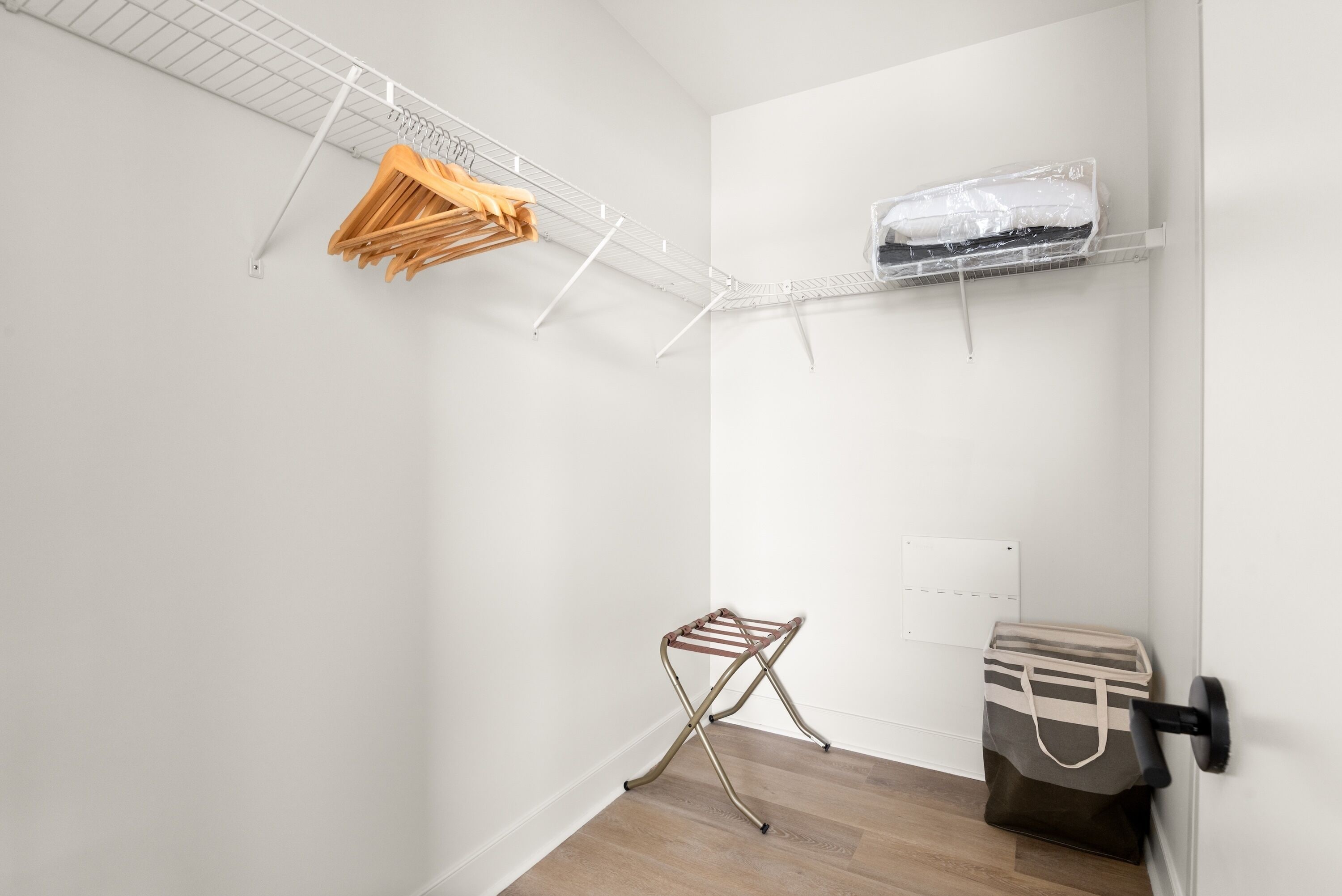 A compact room with a white wall, a wooden floor, a metal drying rack, a small storage unit, and a white ceiling.