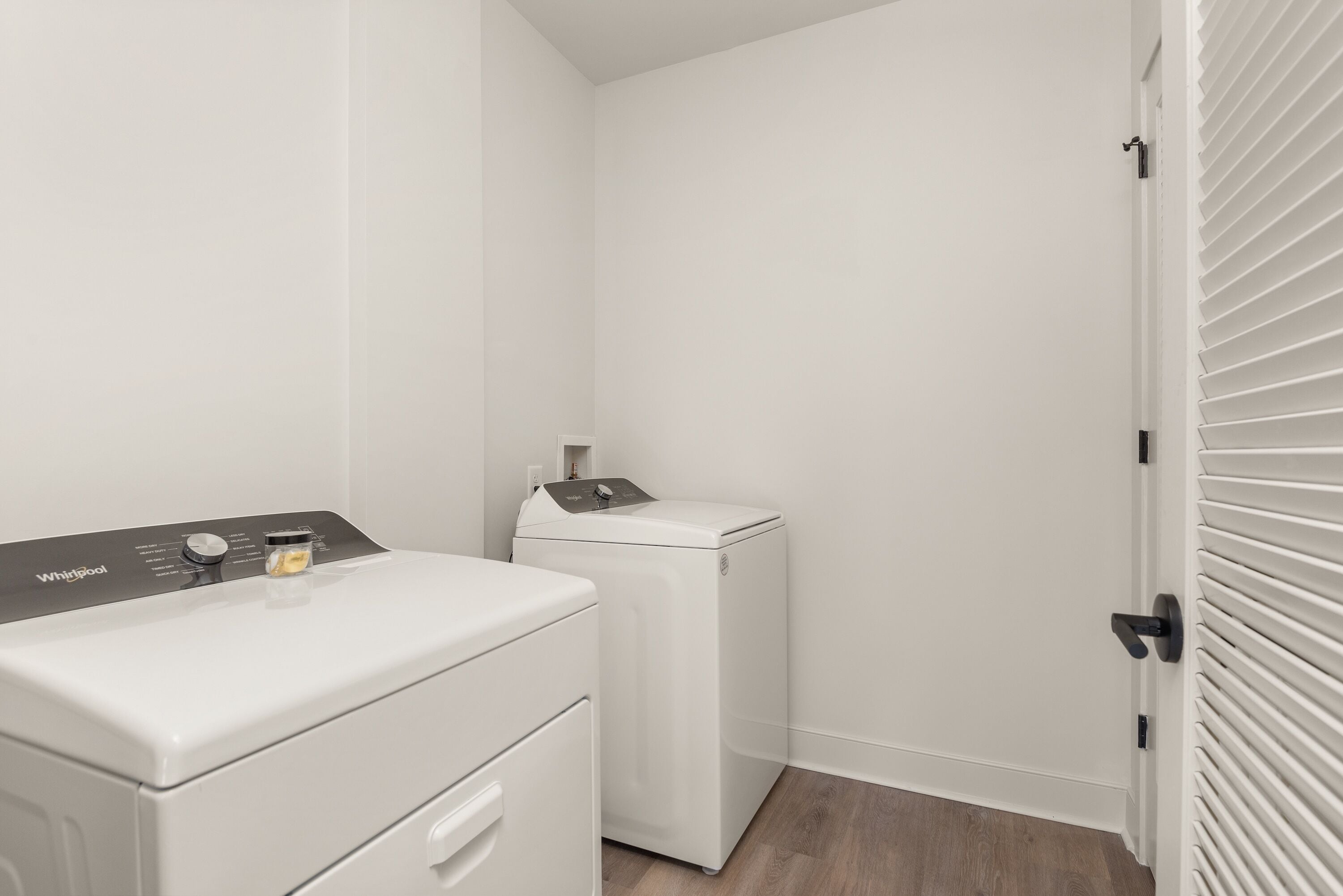 A laundry room with a front-loading washer and dryer, a small cabinet, and a door with blinds.