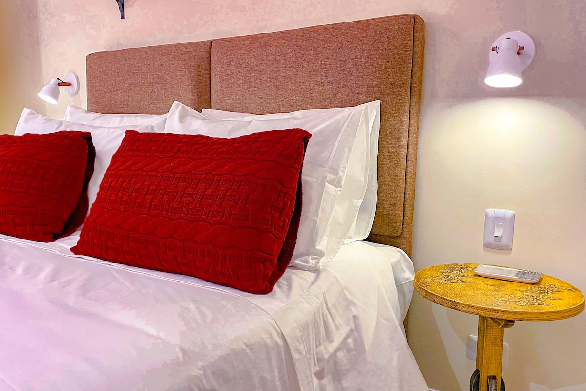 A neatly made bed with white and red bedding, a round wooden side table, and a wall-mounted light fixture.