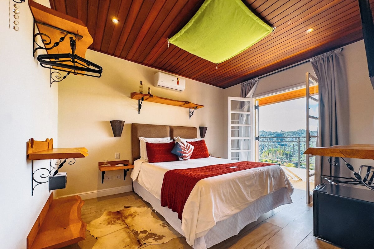 A bedroom with a bed, a wooden floor, a hanging light fixture, a window with curtains, and a view of the outside.