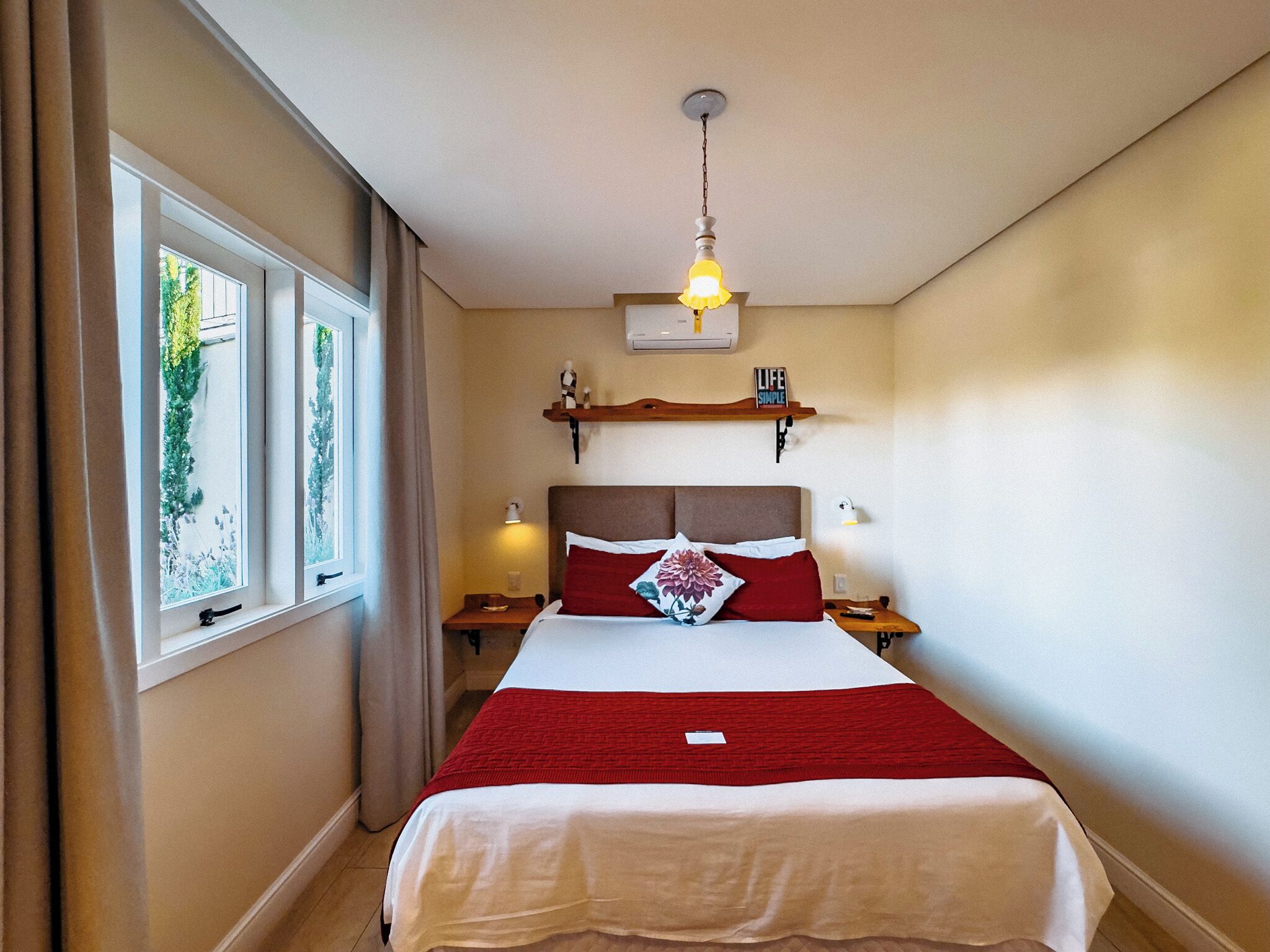 A hotel room with a bed, a window, a ceiling fan, and a wall-mounted shelf with a sign.