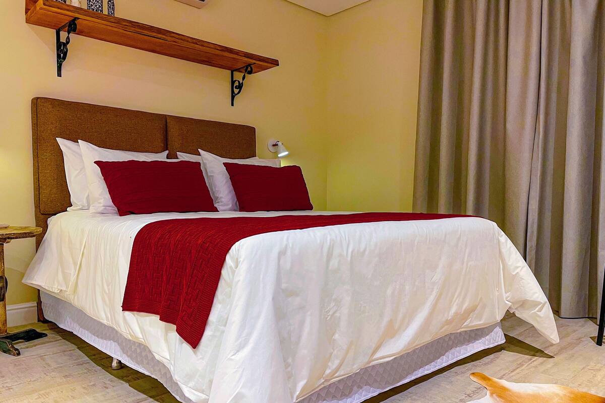 A bed with white and red bedding, a wooden headboard, a nightstand with a lamp, and a large window with curtains.