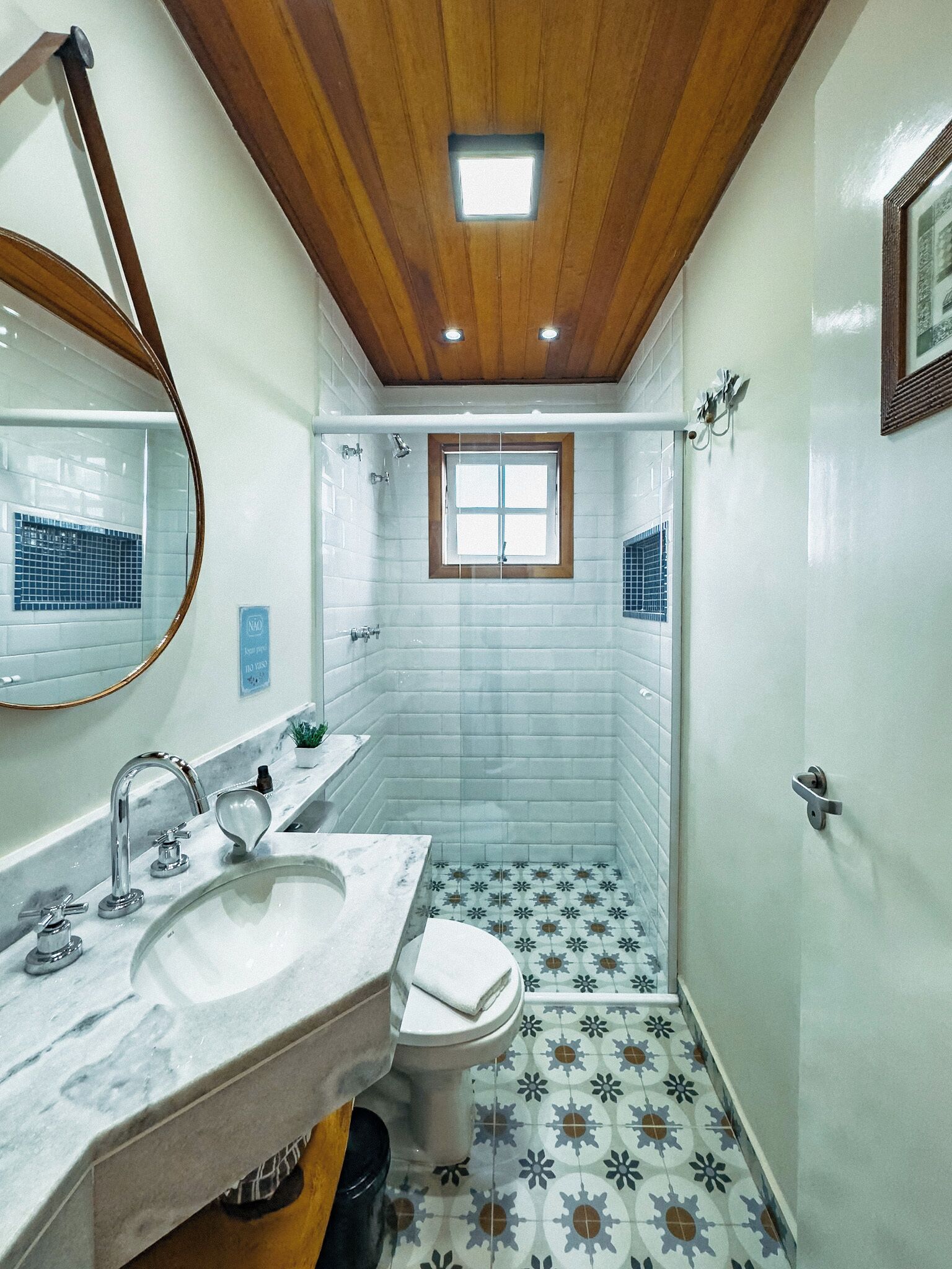 Baño con techo de madera, una ventana, lavabo, inodoro y baldosas de suelo con diseño.