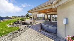 A covered patio area with a hot tub, stone pathway, and a view of a green landscape. - Canyon Rim Retreat (Filer)