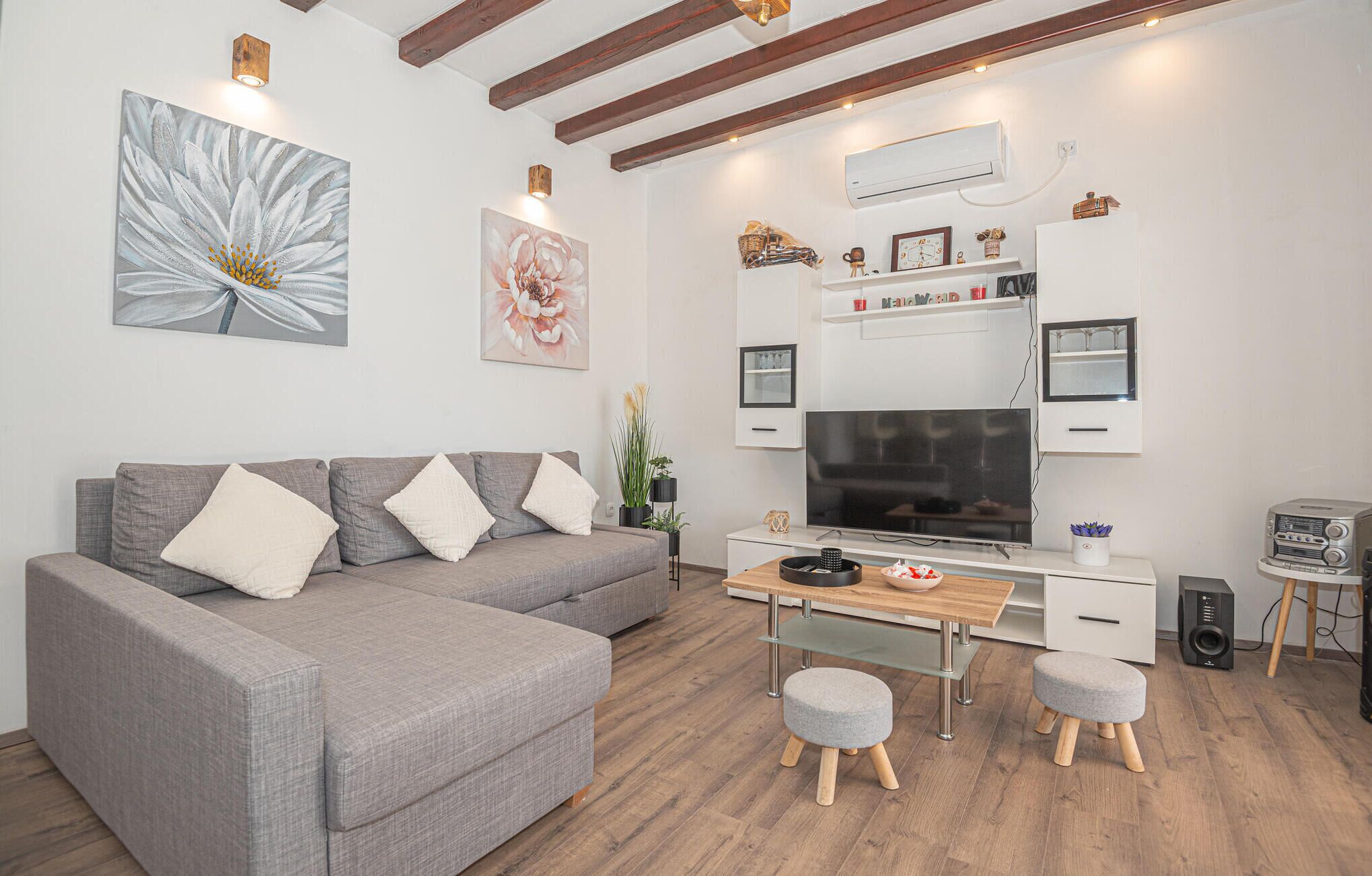 A modern living room with a grey sofa, a wooden desk, a flat-screen TV, and a small kitchenette area.