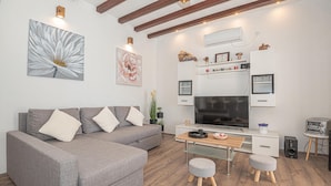 Un salon moderne avec un canapé gris, un bureau en bois, un téléviseur à écran plat et un petit coin cuisine.