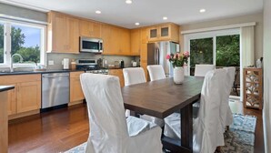 A modern kitchen with wooden cabinets, stainless steel appliances, and a dining area with white chairs and a wooden table.