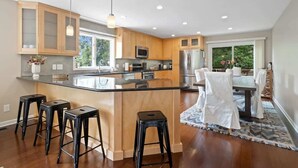 A modern kitchen with wooden cabinets, a central island with stools, stainless steel appliances, and a dining area with white chairs.