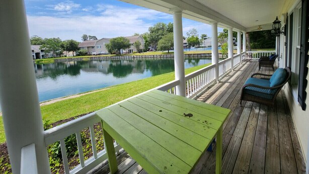 A covered porch with a green table and wicker chairs, overlooking a calm lake and residential buildings. - F3, close to front heated pool (St. Simons Island)