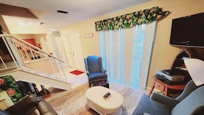 A living room with a staircase, a black leather couch, a grey armchair, a white ottoman, and a television on a wooden stand. - F3, close to front heated pool (St. Simons Island)