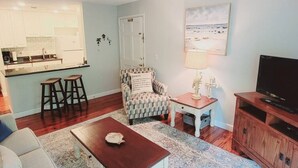 A living room with a kitchen in the background, a patterned armchair, a small table with a decorative item, a television on a wooden stand, and a painting on the wall. - E10 Beautifully decorated 2BR, 2 BA island condo (St. Simons Island)