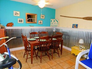 A dining area with a wooden table and chairs, a ceiling fan, and a wall with framed pictures and a sign that reads "ON BEACH TIME".