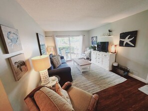 A living room with a sofa, a coffee table, a lamp, a TV, and a fireplace. There is a window with curtains, a rug, and artwork on the walls. - S3, comfy, close to pickleball (St. Simons Island)