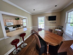 A spacious dining area with a long wooden table and six chairs, a kitchen with modern appliances, and large windows providing natural light. - S3, comfy, close to pickleball (St. Simons Island)