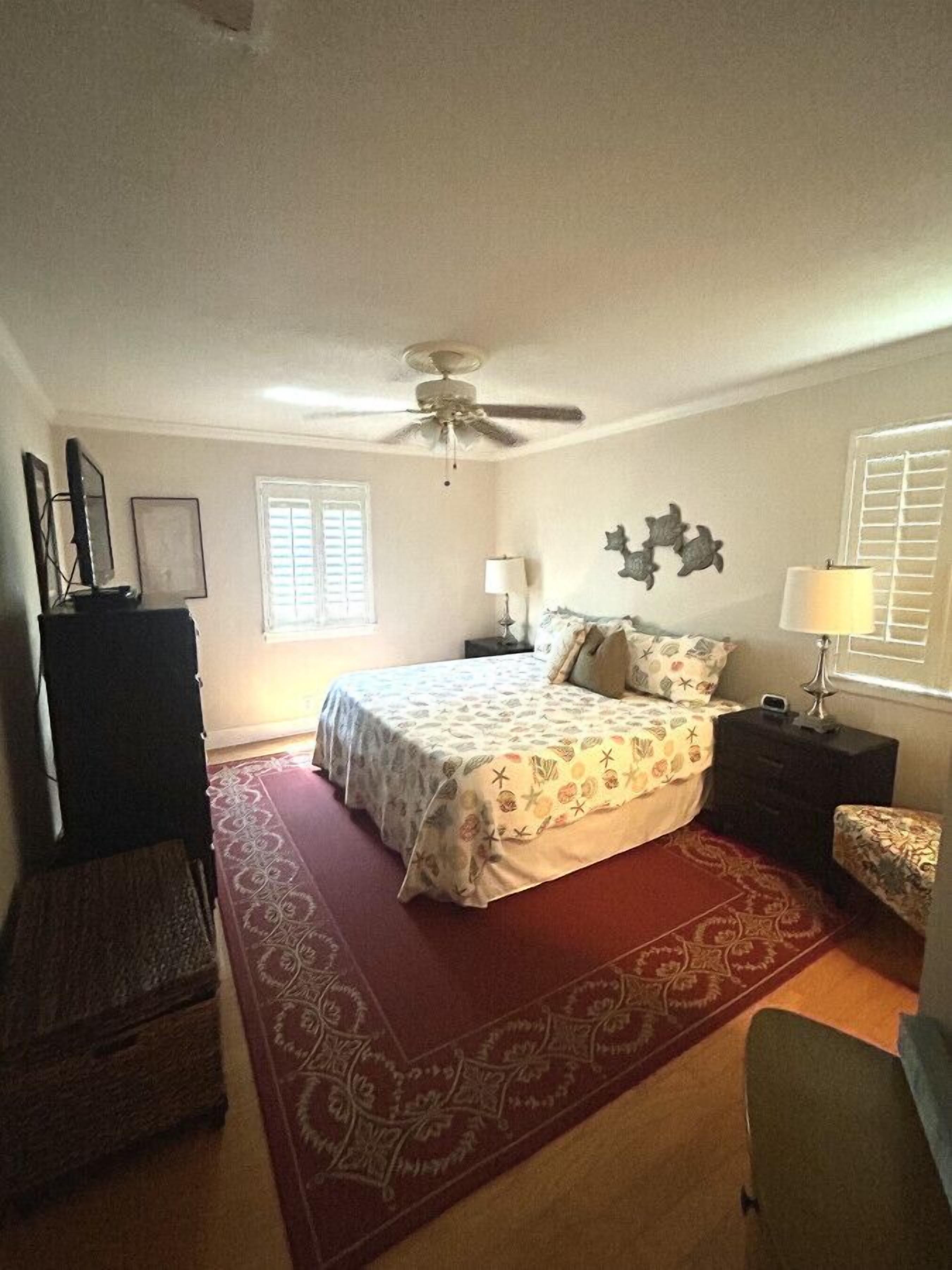 Une chambre à coucher comprenant un lit, un ventilateur de plafond, une commode, une table de chevet, une lampe, une chaise et une fenêtre.