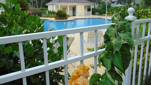 Une piscine entourée d’une clôture blanche, une maison en arrière-plan et un grand parasol.