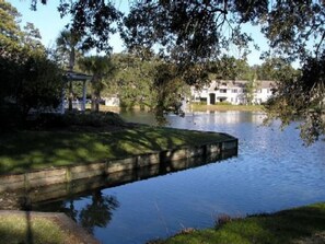 A calm lake with a wooden dock extending into the water, surrounded by trees and a building in the background.