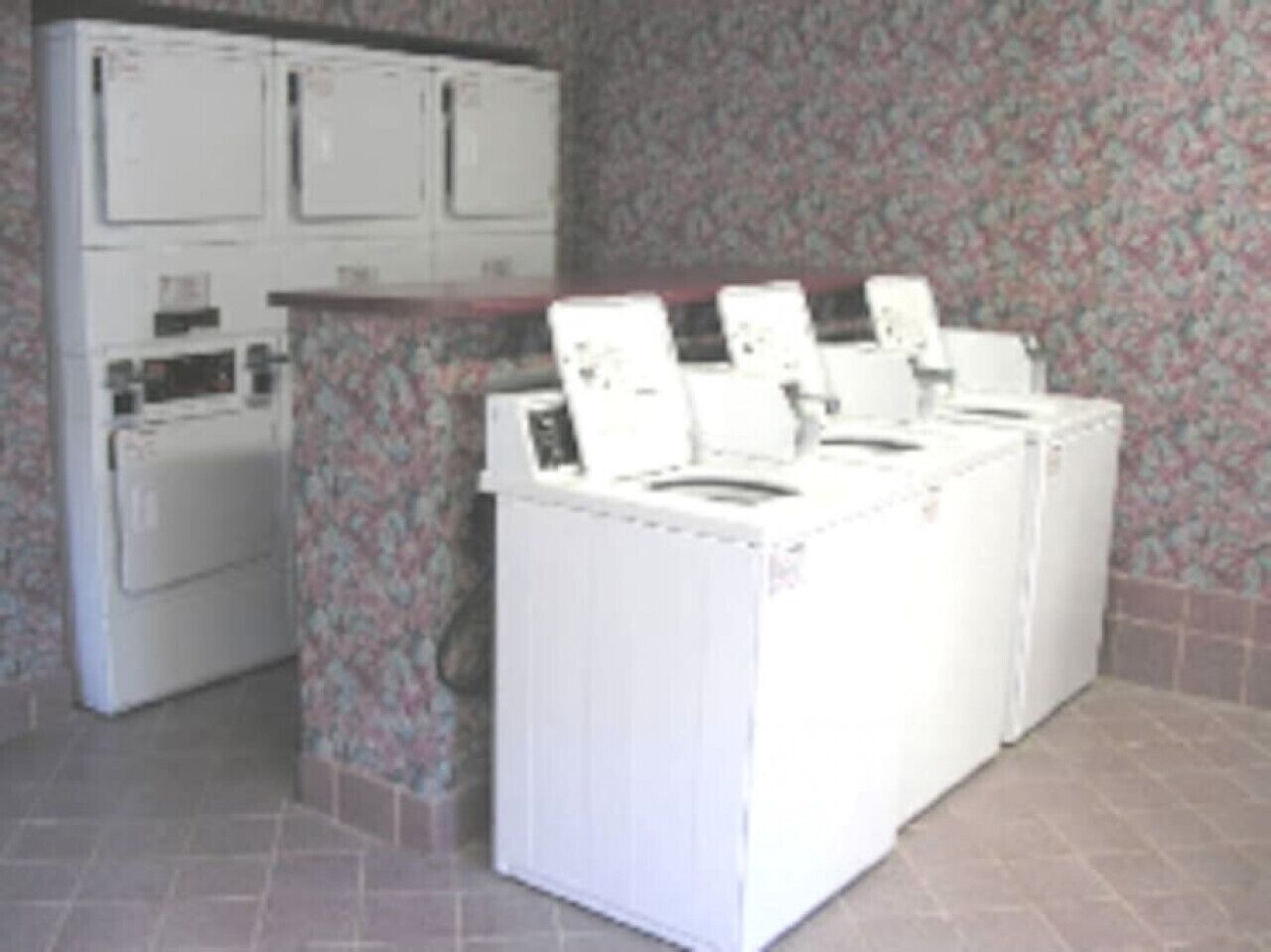 A laundry area with four washing machines and a dryer, tiled walls, and a patterned wallpaper.