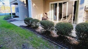Ein Wohngebäude mit überdachter Veranda, einer Schiebetür und einem kleinen Gartenbereich mit geschnittenen Büschen.