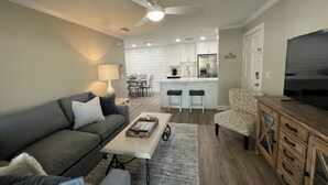 A modern living room with a grey sofa, a coffee table, and a patterned armchair. A kitchen area with white cabinets and a dining set is visible in the background. - H6 Complete renovation, 2nd floor 2 BD/2 bath, close to pool, village and beach (St.  Simons Island)