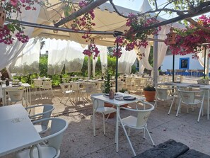 An outdoor patio with white tables and chairs, covered by a white canopy, and decorated with hanging pink flowers.