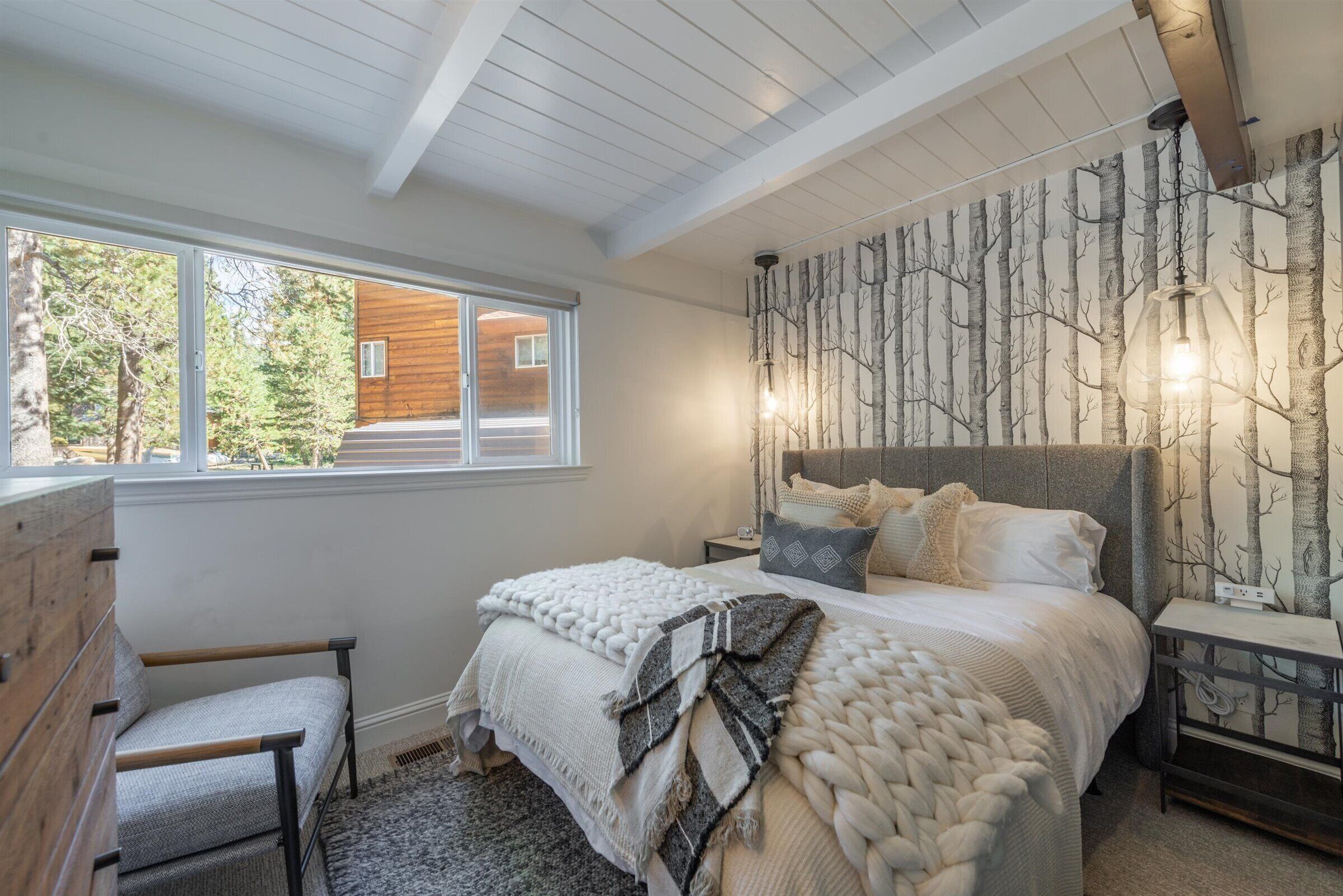 A bedroom with a large bed, a window, a wall with a tree pattern, and a wooden dresser.