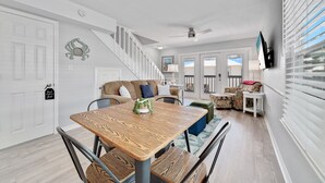 A spacious living area with a wooden dining table, comfortable seating, a staircase, and a television mounted on the wall. - LAGOON VIEWS, OUTDOOR POOL, WALK TO DINING AND SHOPPING! (Gulf Shores)