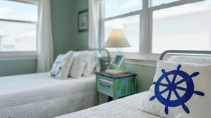 A bedroom with two beds, a window with a view, a bedside table with a lamp, and a pillow with a ship's wheel design. - LAGOON VIEWS, OUTDOOR POOL, WALK TO DINING AND SHOPPING! (Gulf Shores)