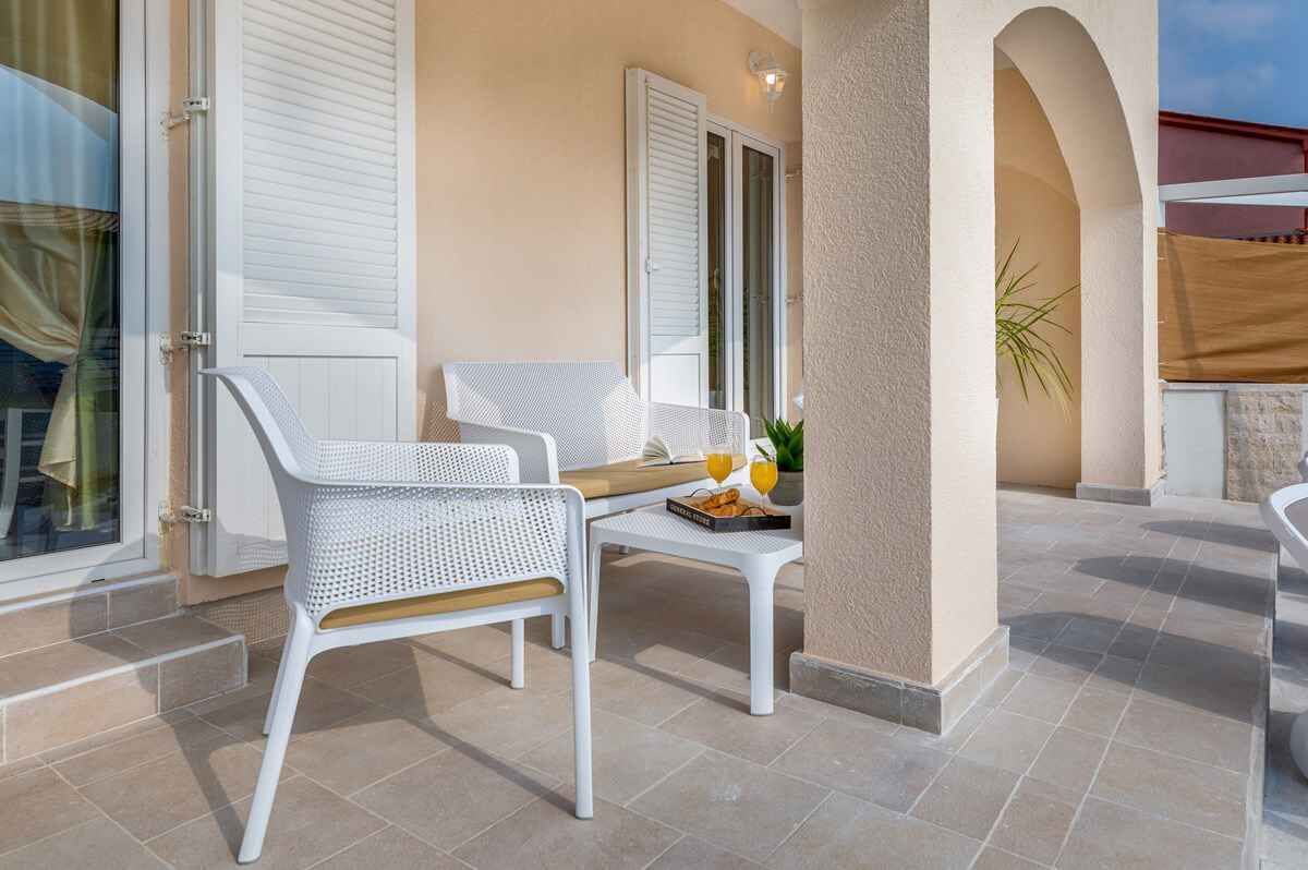 A sunlit outdoor seating area with white chairs and a table, a door with shutters, and a potted plant.