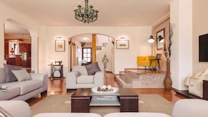 A spacious living room with a sofa set, a coffee table, and a chandelier. There is a staircase leading to an upper level.