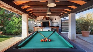 An outdoor pool area with a pool table, wooden beams, and a kitchen area under a covered patio.