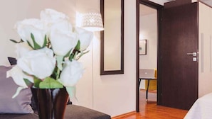 A hotel room with a bed, a vase of white flowers, a lamp, and a door leading to another room.